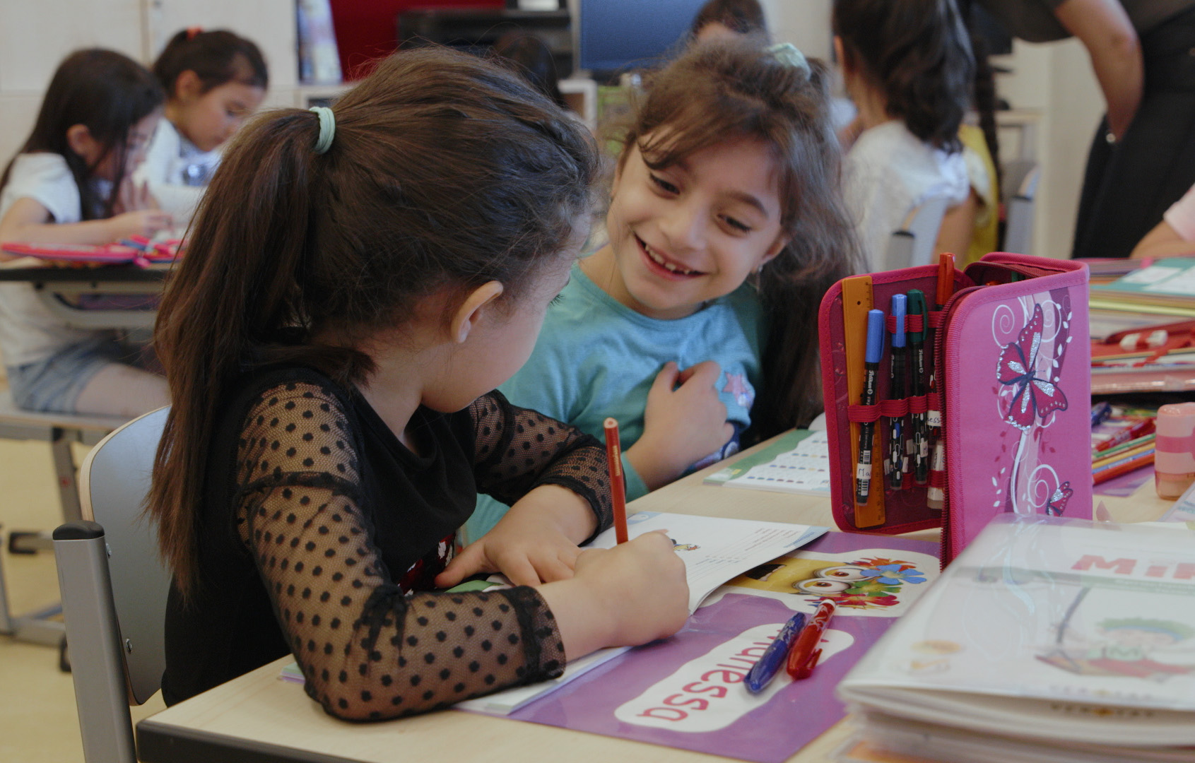 Zwei Kinder sitzen gemeinsam an einem Schultisch und schauen sich gegenseitig an