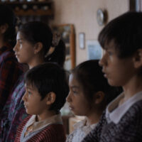 Die fünf Geschwister leben gemeinsam mit ihrem Vater und der Großmutter in einem Haus in Brasilien. Die Mutter ist verschwunden und das Einzige, was von ihr zurückgeblieben ist, sind Videoaufzeichnungen, auf denen sie gemeinsam mit den Kindern zu sehen ist und in dessen Rückblicken es phasenweise so wirkt, als sei hier die Welt noch in Ordnung gewesen. Als eines Morgens für jedes Kind ein paar „neue“ Schuhe vor der Tür standen, vermutet der Vater, das diese vermutlich von der Mutter in der Nacht gebracht wurden. Er macht sich kurzer Hand auf den Weg, um seine Partnerin zu suchen. Allerdings kehrt auch er nicht zurück und so sind die Kinder mehr oder weniger auf sich alleine gestellt. Das Zusammenleben mit der Großmutter gestaltet sich recht speziell. Sie glaubt fest daran, das der Teufel kommen wird, wenn man nicht gut genug aufpasst oder gewisse Regeln nicht einhält. Dieser starke Glaube spiegelt sich […]